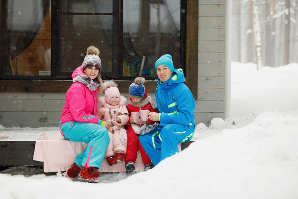 A család téli ruhában ül a verandán és teát vagy kávét iszik. Vidéki ház egy havas tűlevelű erdőben. Meleg zakók, kötött sapkák, poharak. Családi boldogság, értékek és hagyományok. - Fotó, kép