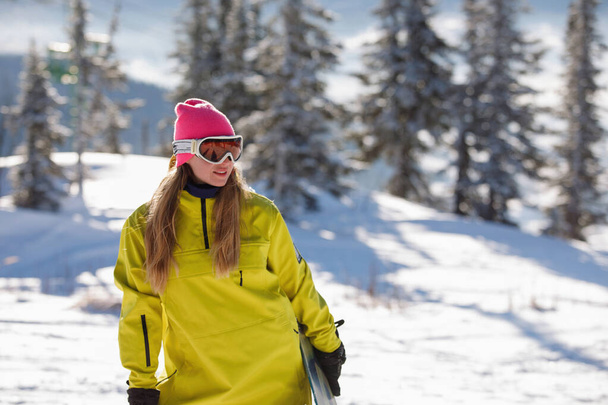 Сноубордистка в лыжной одежде позирует с доской на фоне синих гор. Желтая куртка с капюшоном, вязаная шляпа, лыжные очки. Здоровый образ жизни. Спортивная концепция. Селективный фокус. - Фото, изображение
