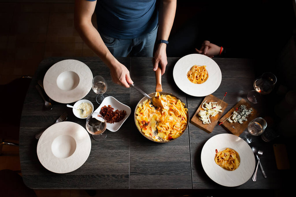 Man sets the table, homemade pasta, bacon, feta and tomatoes, cheeses with blue mold, wine. Deep plates for pasta. - 写真・画像