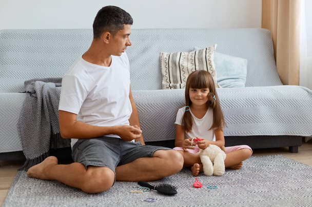 Indoor shot of handsome man listening his daughter attetively, family wearing ccasual style clothing sitting on floor near sofa, spending time for conversation. - Foto, Imagen