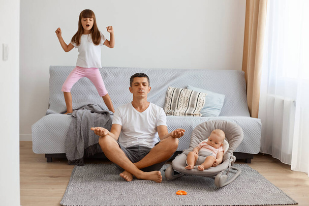 Young adult man sitting on floor near sofa with his little kid, wearing white t shirt and jeans short, trying to relax with yoga while the eldest daughter sings loudly. - Foto, afbeelding