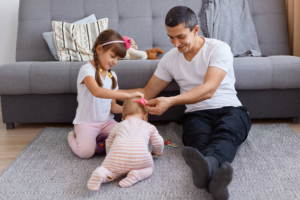 Indoor shot of Caucasian handsome brunette father playing with his daughters while sitting on floor near sofa, wearing white t shirt and trousers, expressing happiness. - 写真・画像