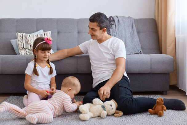 Young adult man with pleasant appearance sitting on floor near sofa and playing, with his little kids, looking at children with love.wearing white t shirt. - 写真・画像