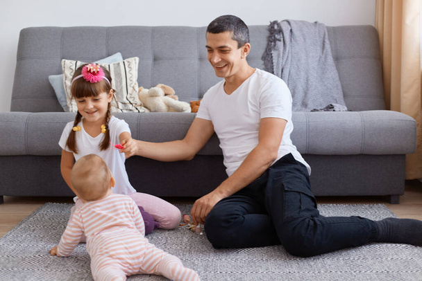 Smiling attractive young adult man sitting on floor near sofa and playing with his little kids, wearing white t shirt and trousers, looking at his crawling baby kid and laughing, - Photo, image