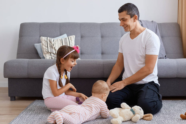 Indoor shot of attractive young adult father sitting on floor near sofa and playing with his little kids, expressing positive emotions and happiness, parenthood anfd childhood. - Фото, изображение