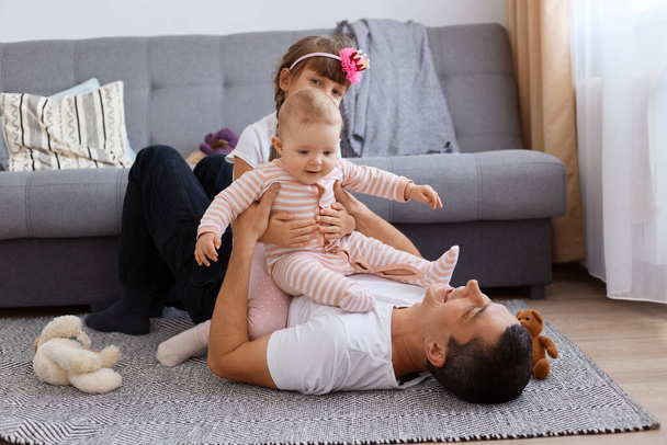 Extremely happy young adult man lying on floor near sofa and playing with his little kids, wearing white t shirt and trousers, children sitting on father, family enjoying spending time together. - 写真・画像