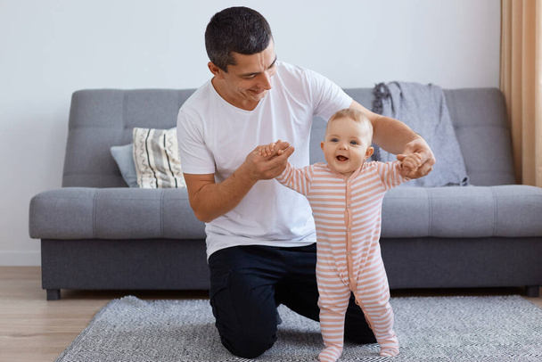 Indoor shot of happy smiling brunette man wearing white casual style t shirt holding daughter's hands, teaching her to go, expressing positive emotions and happiness. - Фото, зображення