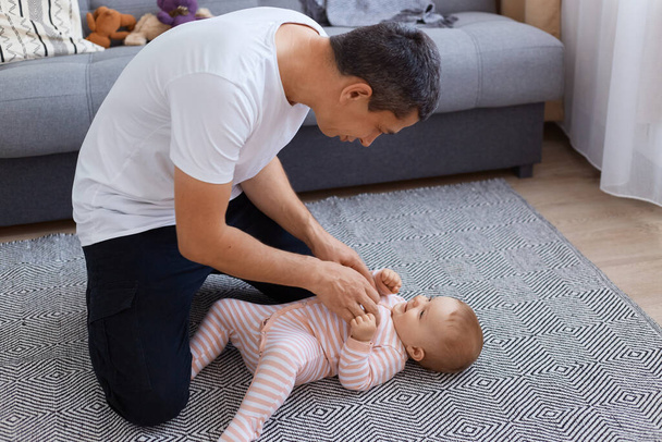 Indoor shot of playful brunette man wearing white t shirt playing with his little infant daughter at home, cute child in sleeper lying on floor and looking at father. - Foto, immagini