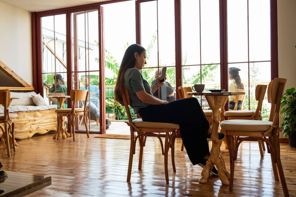 Młoda kobieta w luźnej koszuli siedzi przy stole w kawiarni i sprawdzając posłańca podczas korzystania ze smartfonu.Pełne body shot we wnętrzu kawiarni  - Zdjęcie, obraz