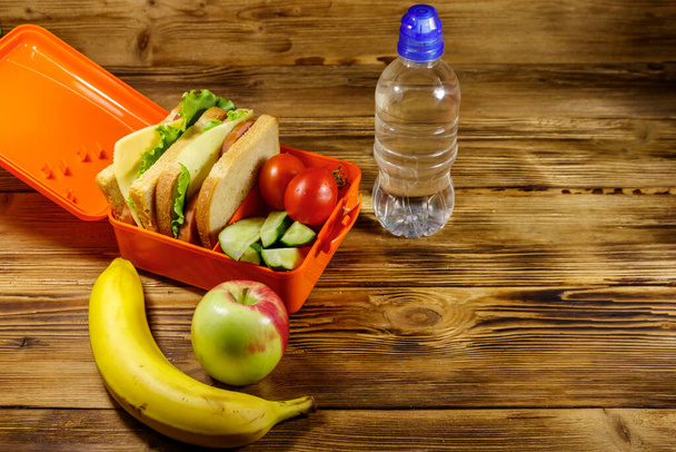 Bottle of water, apple, banana and lunch box with sandwiches and fresh vegetables on a wooden table - Photo, Image