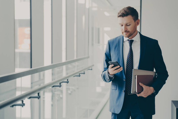 Сосредоточенный бизнес-консультант одет официально держа в руках мобильный телефон, читая новости онлайн в офисе - Фото, изображение