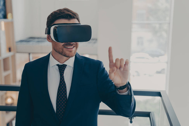 Улыбающийся успешный бизнесмен в костюме, стоящий в офисном интерьере и использующий очки VR для работы - Фото, изображение