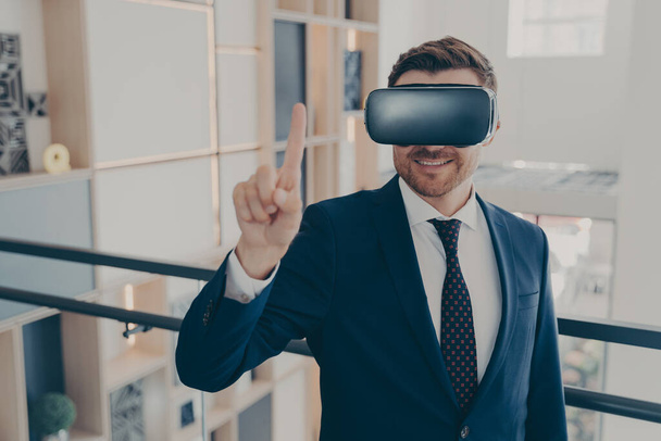 Young cheerful businessman in formal suit and VR headset goggles standing in company lobby - Valokuva, kuva