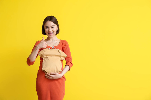 Νεαρή έγκυος γυναίκα με κοστούμι του μωρού δείχνει αντίχειρα-up σε κίτρινο φόντο - Φωτογραφία, εικόνα