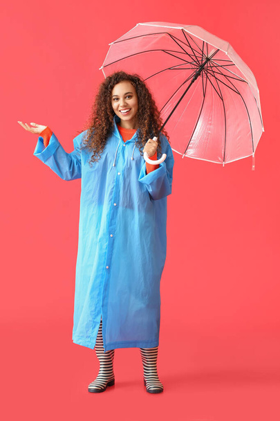 Young African-American woman wearing blue raincoat and gumboots with umbrella on red background - Foto, Imagen