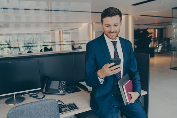 Sebevědomý usmívající se podnikatel pomocí smartphone a usmívat se při stání v úřadu - Fotografie, Obrázek