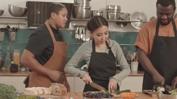 Medium van jonge Aziatische vrouw en Afro-Amerikaanse man dragen schorten, snijden komkommers op snijplanken met messen, kortharige vrouwelijke chef-kok kijken naar hen en het geven van advies - Video