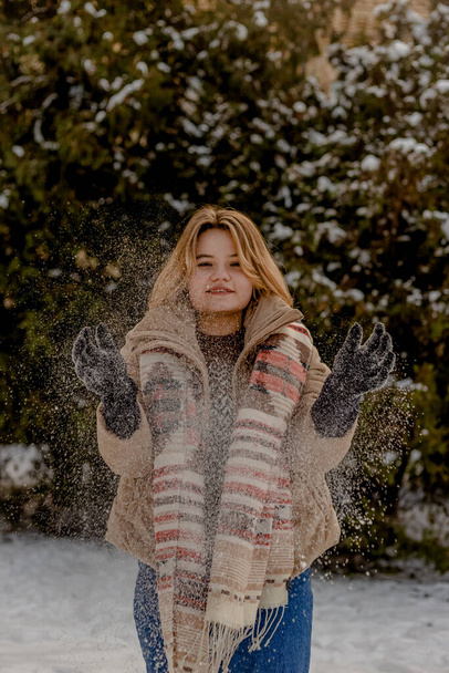 Ξανθιά νεαρή γυναίκα με μπεζ σακάκι και μαντήλι ρίχνει χιόνι με τα χέρια της. Ιστορικό χριστουγεννιάτικων δέντρων στο χιόνι. Χειμερινή περίοδος. - Φωτογραφία, εικόνα