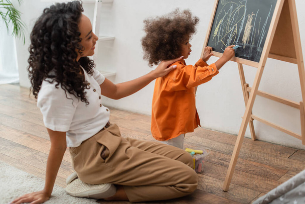 Toddler kid drawing on chalkboard near smiling african american mother at home  - Fotografie, Obrázek