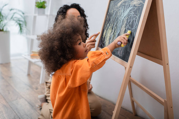 Criança africana americana menina desenho no quadro perto da mãe em casa  - Foto, Imagem