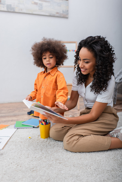 Donna afro-americana positiva che tiene libro vicino a figlia e matite di colore a casa  - Foto, immagini