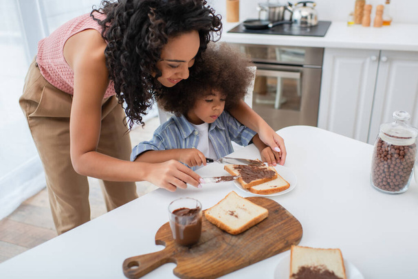 Smiling african american mom spreading chocolate paste on bread near daughter in kitchen  - 写真・画像