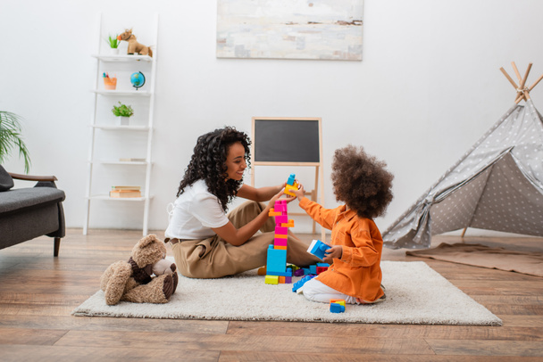 Mosolygó afro-amerikai anya játszik építőkockák gyermek közelében Teddy maci otthon  - Fotó, kép