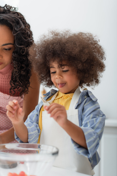 Criança afro-americana criança em avental de pé perto da mãe e tigela na cozinha  - Foto, Imagem