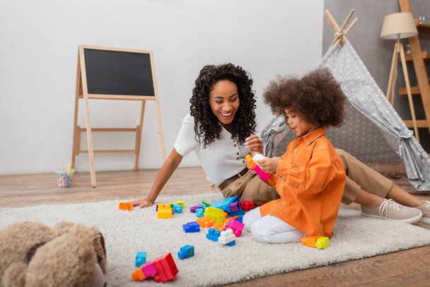 Positive african american mother sitting near kid with building blocks at home  - Photo, Image