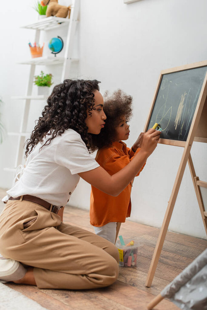 Young african american woman drawing on chalkboard with daughter at home  - Foto, Imagem