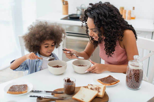 Mère et fille afro-américaine positive assise près des céréales et de la pâte de chocolat pendant le petit déjeuner à la maison  - Photo, image