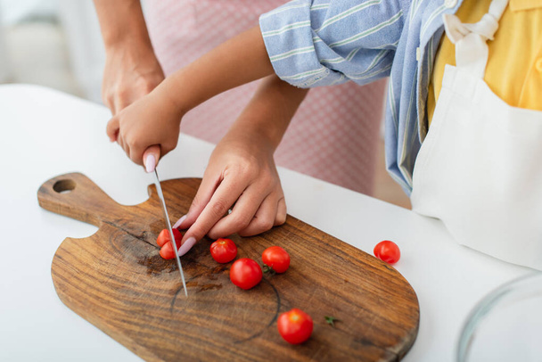 Cropped view of african american kid holding hand of mother with knife cutting cherry tomatoes in kitchen  - Photo, Image