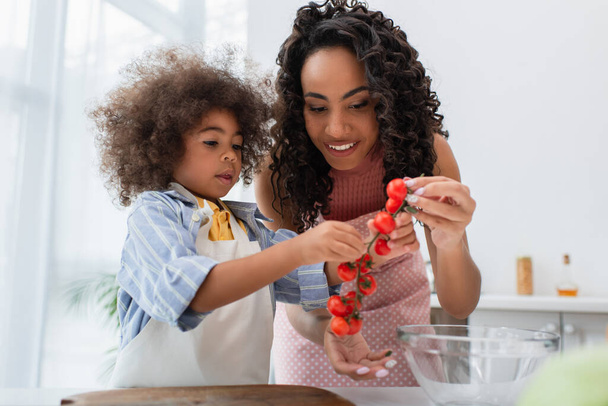 Pozitif Afro-Amerikan annesi ve çocuğu ellerinde kiraz domatesleri ile mutfakta kesim tahtasının yanında  - Fotoğraf, Görsel