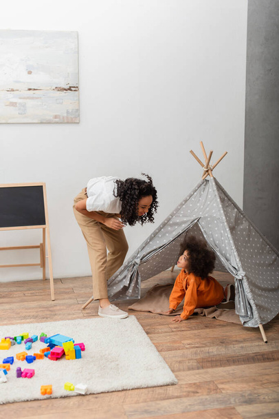 African american mom looking at toddler kid in teepee at home  - Photo, image