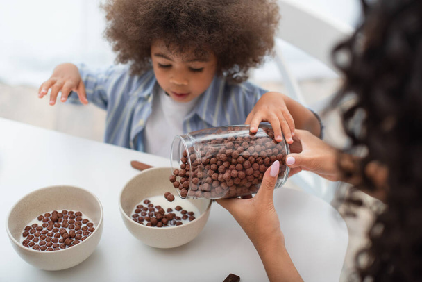 Blurred woman pouring cereal in bowls with milk near african american daughter  - Foto, Imagen