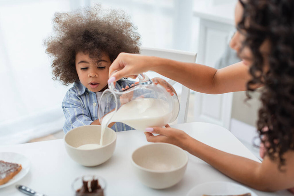 Mère afro-américaine floue versant du lait dans des bols près de la fille dans la cuisine  - Photo, image