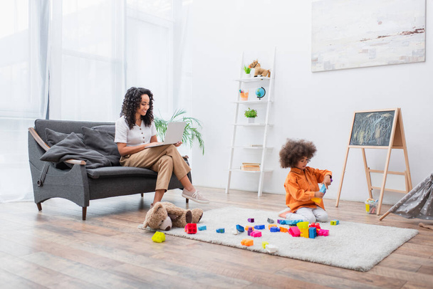 Smiling woman using laptop on couch while daughter playing building blocks at home  - Foto, immagini