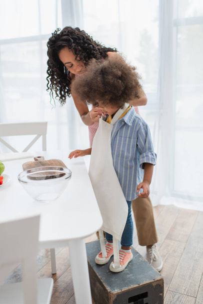 African american woman wearing apron on daughter near bowl and chopping board in kitchen  - Foto, Imagem
