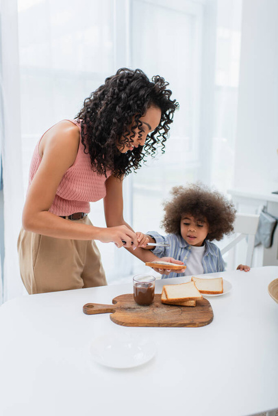 African american woman holding bread and knife near daughter during breakfast at home  - 写真・画像