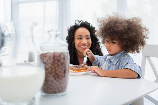 Criança afro-americana criança espalhando pasta de chocolate no pão perto mãe turva na cozinha  - Foto, Imagem
