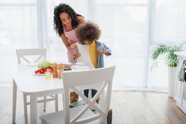 Afro americana mujer usando delantal en hija cerca de verduras en cocina  - Foto, imagen