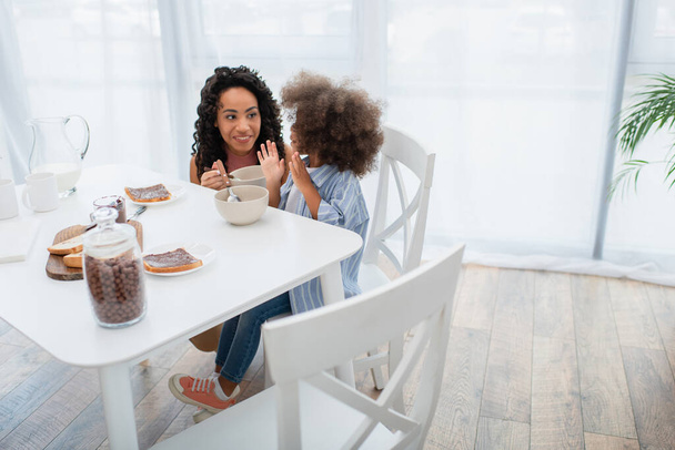 Smiling african american woman holding bowl near daughter and breakfast in kitchen  - Photo, image