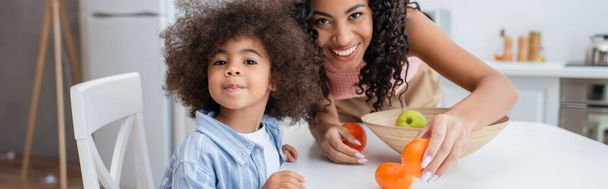 Positive african american woman holding orange near daughter looking at camera in kitchen, banner  - Valokuva, kuva