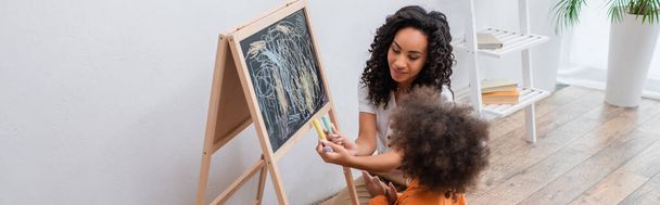 Smiling african american mother holding chalks near daughter and chalkboard at home, banner  - 写真・画像