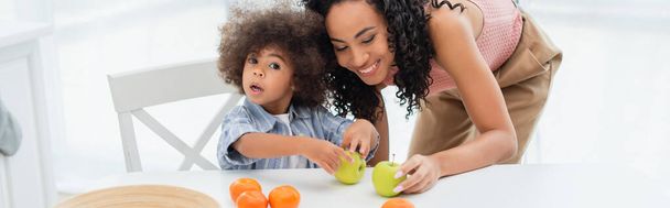 Mamma afroamericana che tiene mela vicino a figlia e arance in cucina, banner  - Foto, immagini