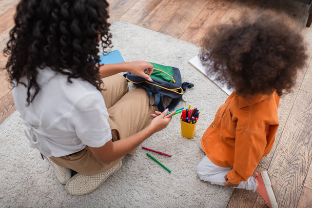 Felülnézetben afro-amerikai anya gazdaság színes ceruzák és hátizsák közelében gyerek otthon  - Fotó, kép