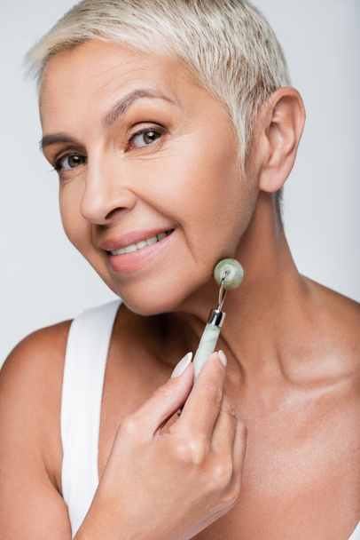 happy senior woman massaging face with jade roller isolated on grey - Fotó, kép
