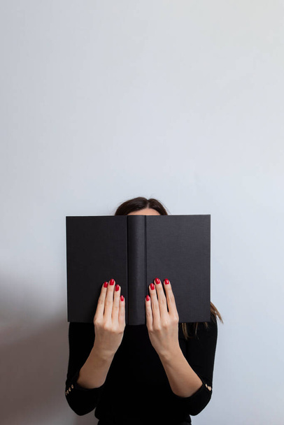 Woman in black clothes holds a black book in her hands that covers her face. White background. Woman reading a book - Fotoğraf, Görsel