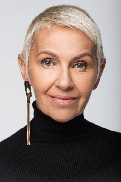 cheerful senior woman with long earring and black turtleneck isolated on grey - Zdjęcie, obraz
