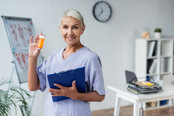cheerful elderly doctor holding bottle with pills and clipboard  - Foto, afbeelding